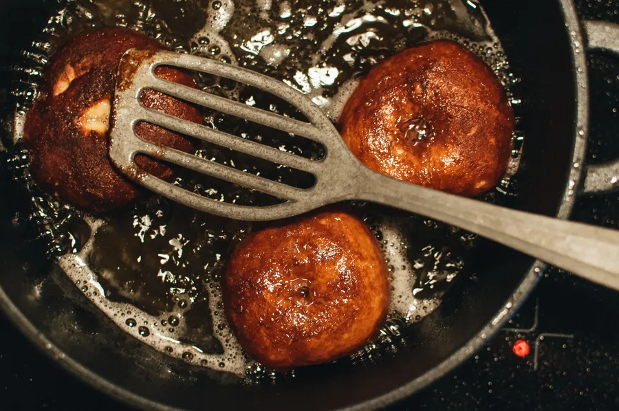 Drei goldbraune Donuts brutzeln in einer Pfanne mit heißem Öl, ein Pfannenwender liegt quer darüber.