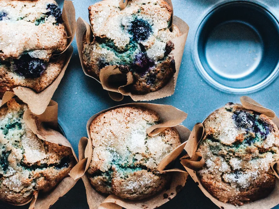 Vier Blaubeermuffins in braunen Papierförmchen auf einem blauen Blech, mit saftigen Beeren und Zuckerkruste auf der goldbraunen Oberfläche.