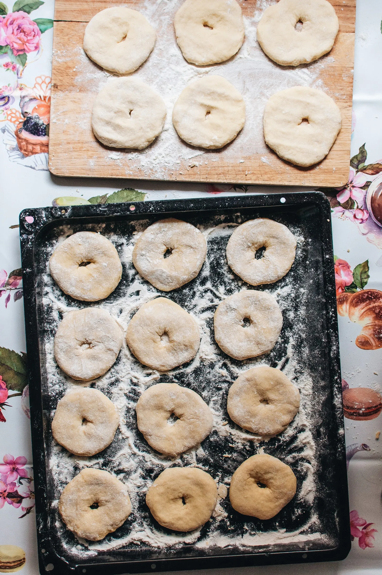 Rohe Donut-Teiglinge auf einem bemehlten Backblech und einem Holzbrett, bereit zum Gehen.
