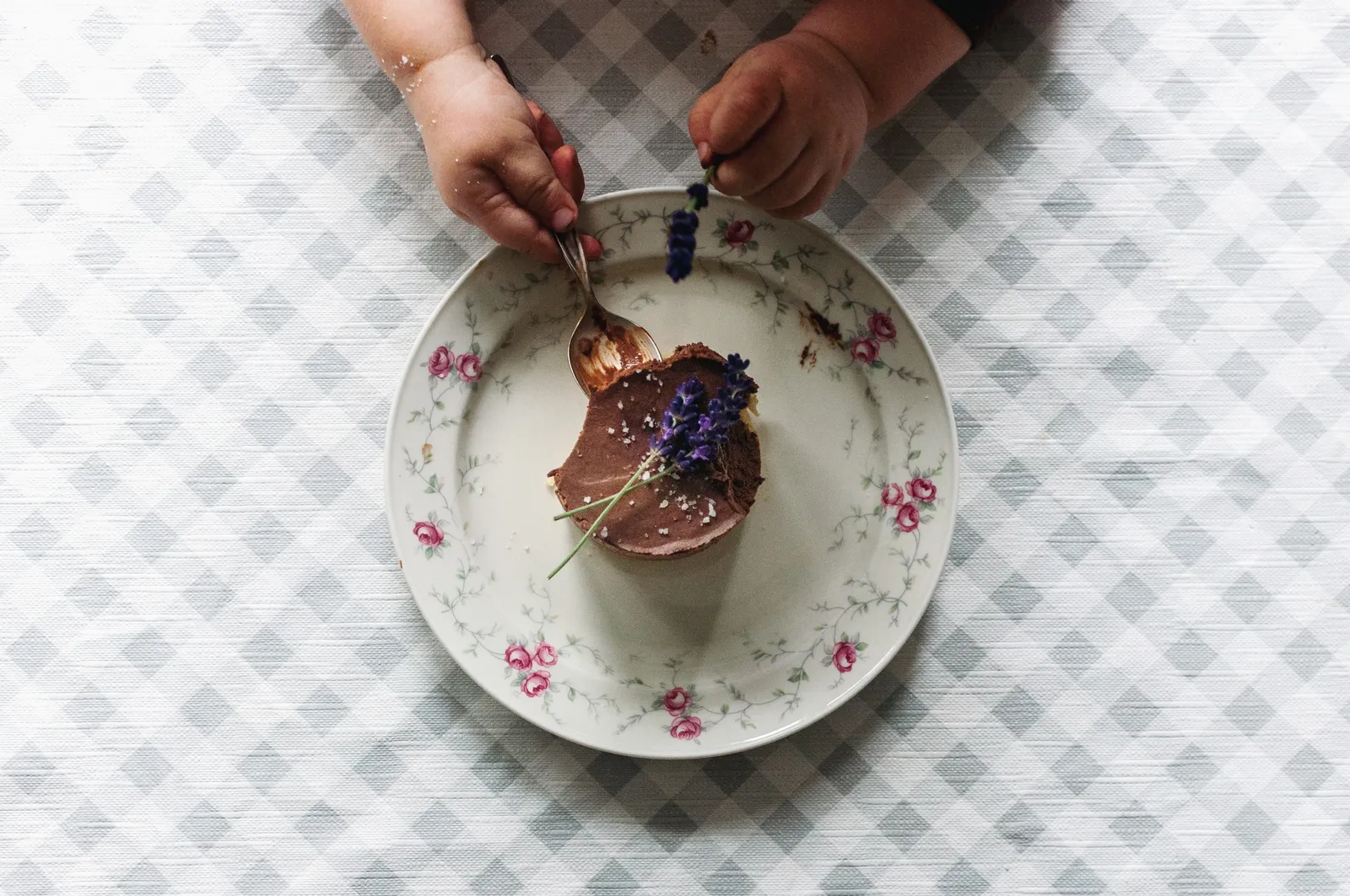 Zwei Hände stechen mit einem Löffel in ein kleines Schokoladenmousse-Törtchen auf einem Blümchenteller, garniert mit einem frischen Lavendelzweig.