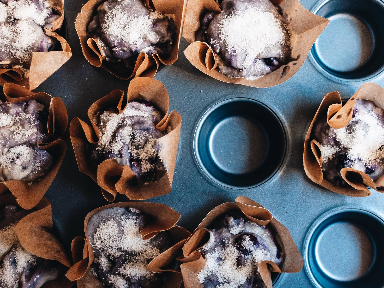 Blaubeermuffins in braunen Papierförmchen in einem blauen Muffinblech, mit Zucker bestreut, von oben fotografiert.