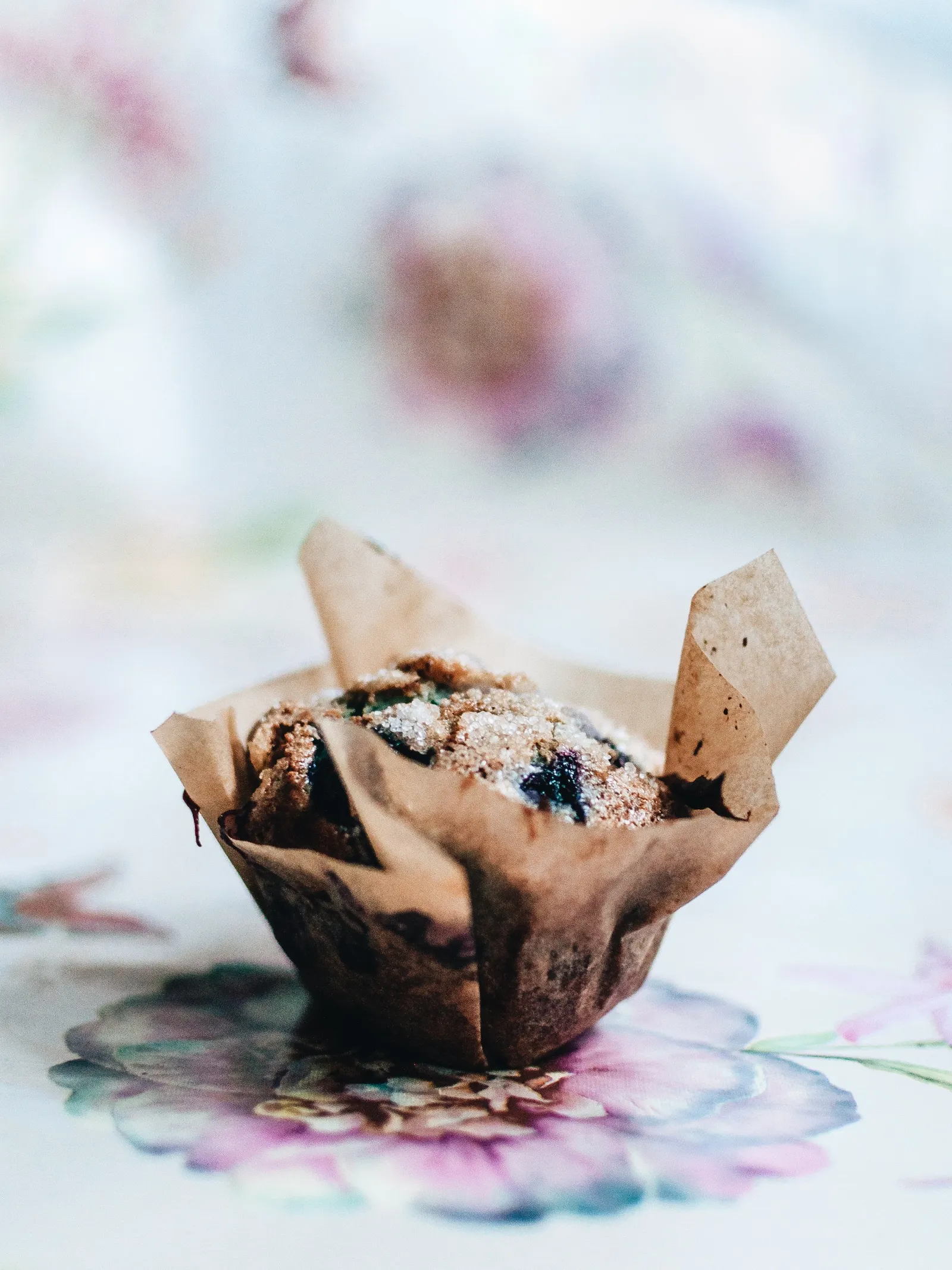 Ein Blueberry-Muffin im zerknitterten Backpapier auf einem Tischtuch mit Blumenmuster, die Oberfläche mit Zuckerkristallen bestreut und dunklen Blaubeeren durchzogen.