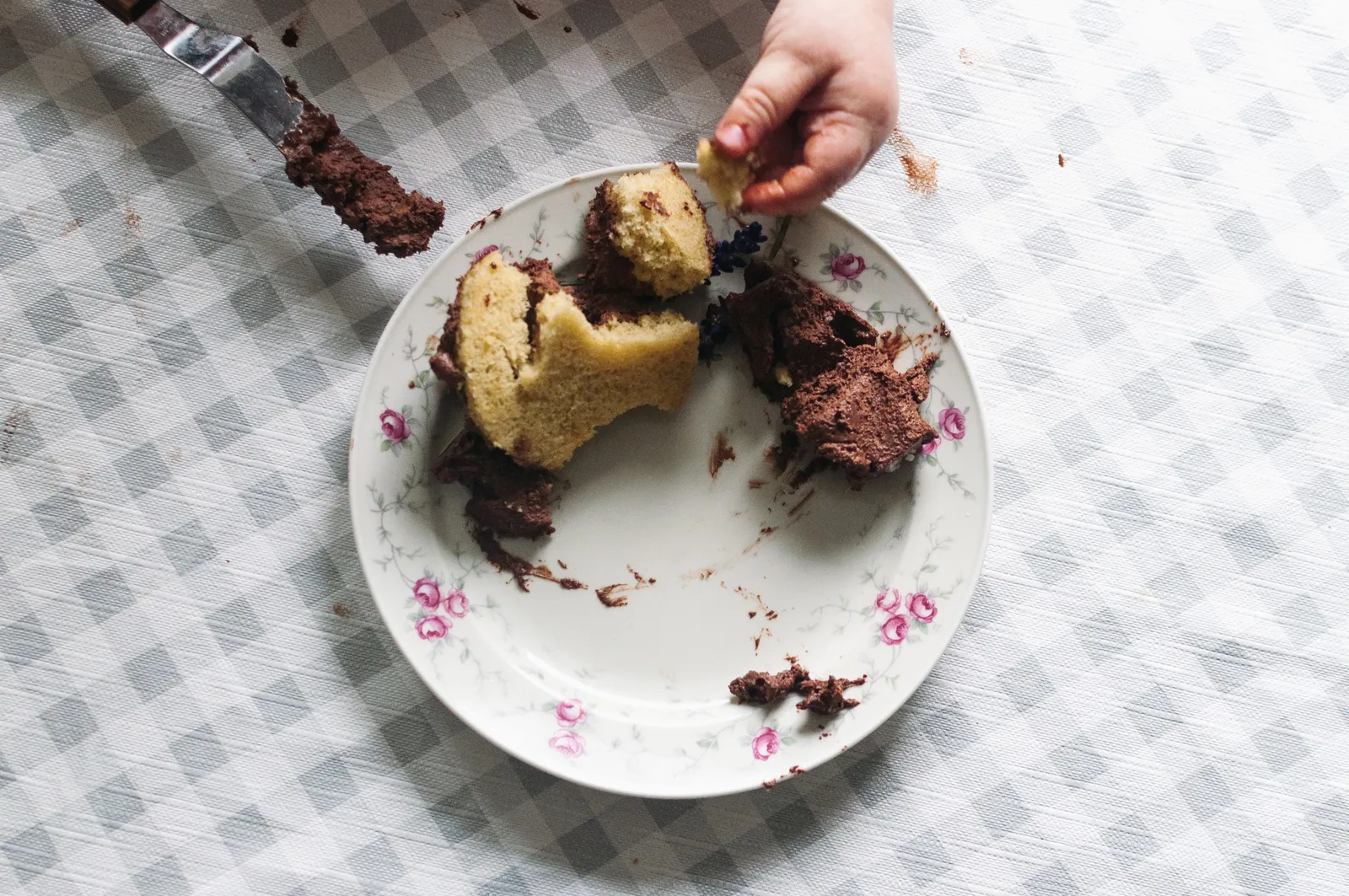 Eine Hand bricht ein angeschnittenes Törtchen mit gelbem Biskuit und dunkler Schokoladenmousse auf einem geblümten Porzellantellerchen auseinander.
