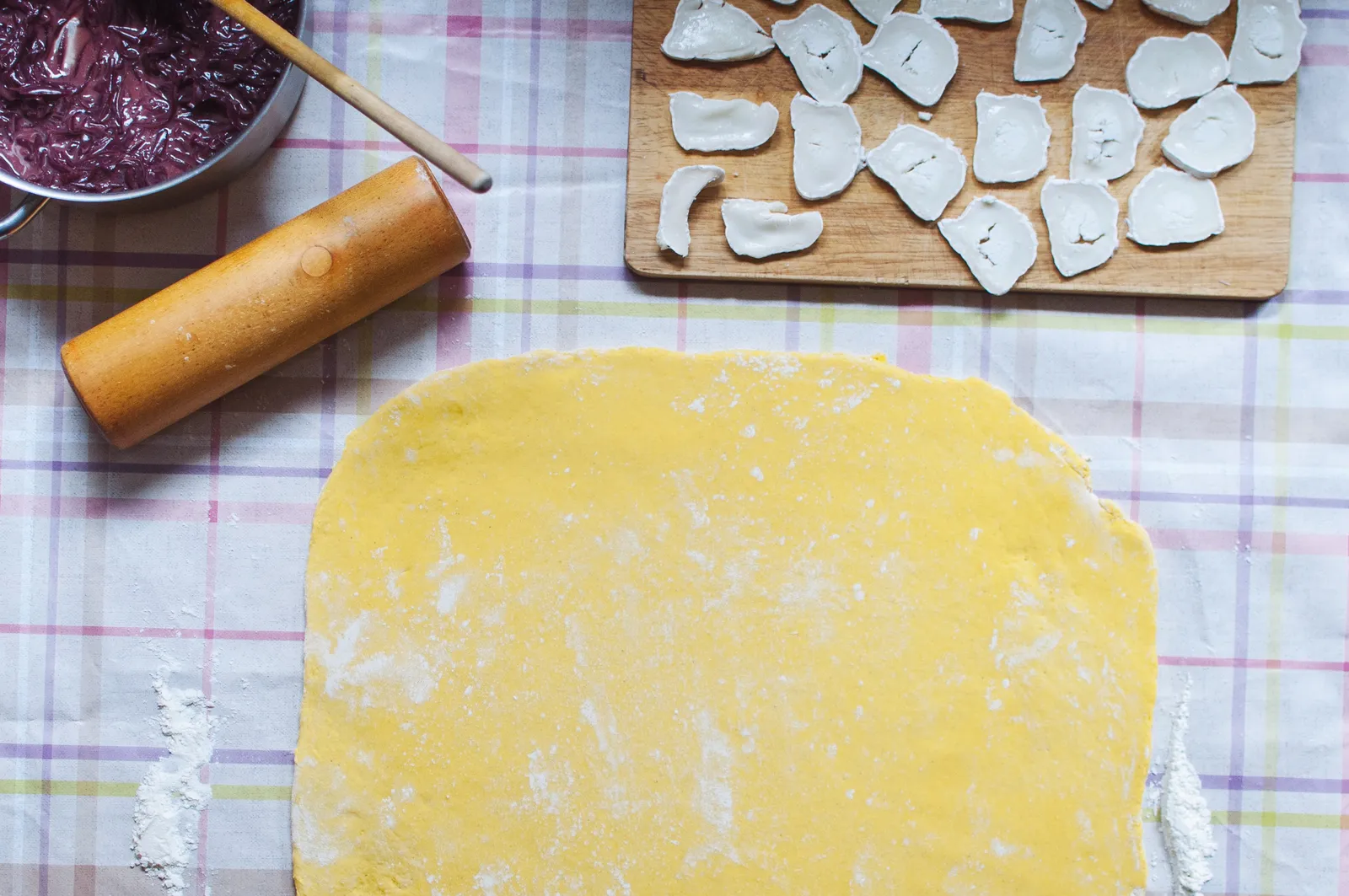 Ausgerollter gelber Mürbeteig auf einem karierten Küchentuch, daneben ein Nudelholz, ein Schälchen mit dunkelroten Rotweinzwiebeln und ein Schneidebrett mit Ziegenkäsestücken.