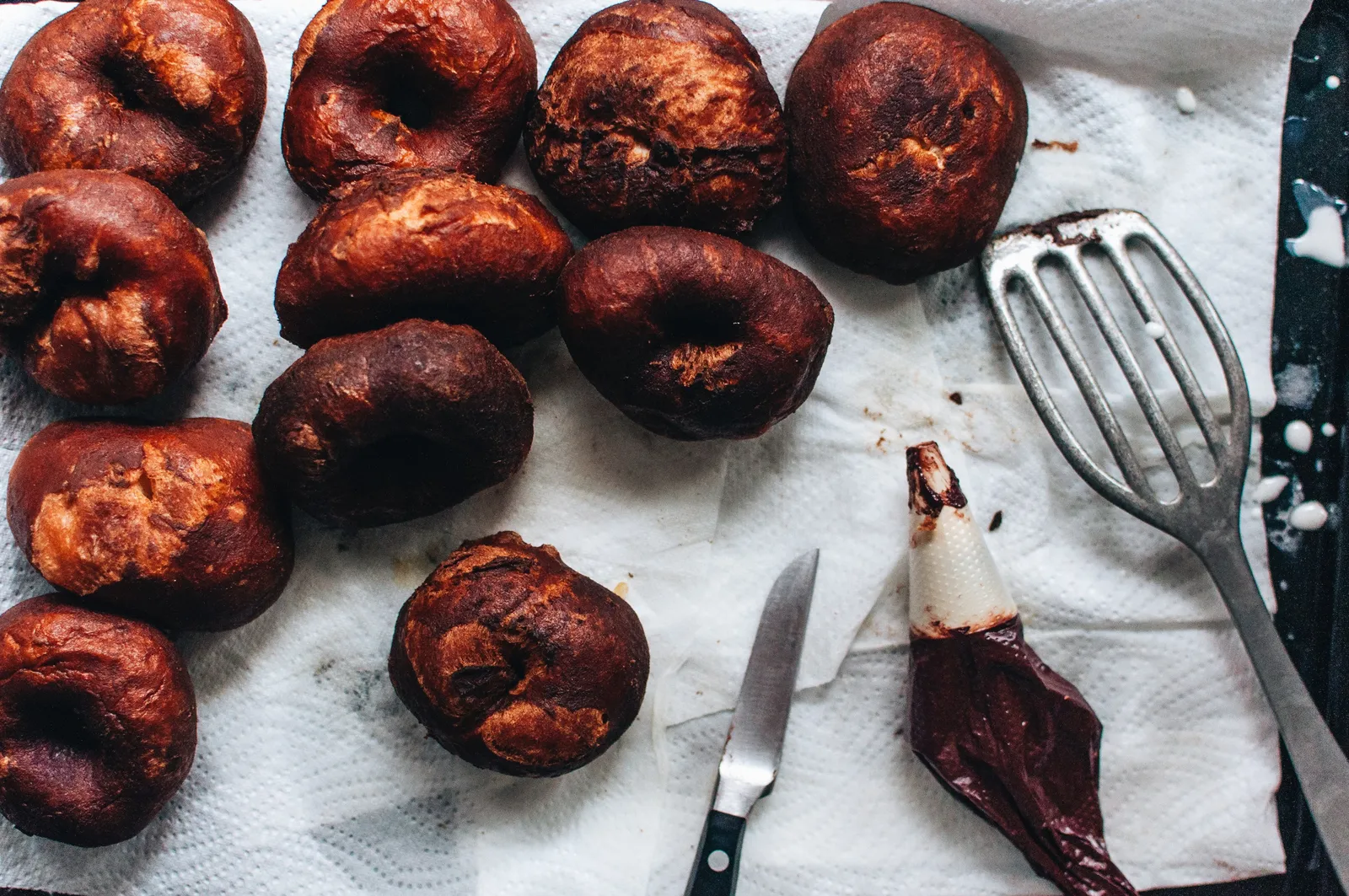 Frisch frittierte Laugendonuts mit tiefdunkler, glänzender Kruste liegen auf Küchenpapier, daneben ein Messer, ein Pfannenwender und ein Spritzbeutel mit weißer Glasur.