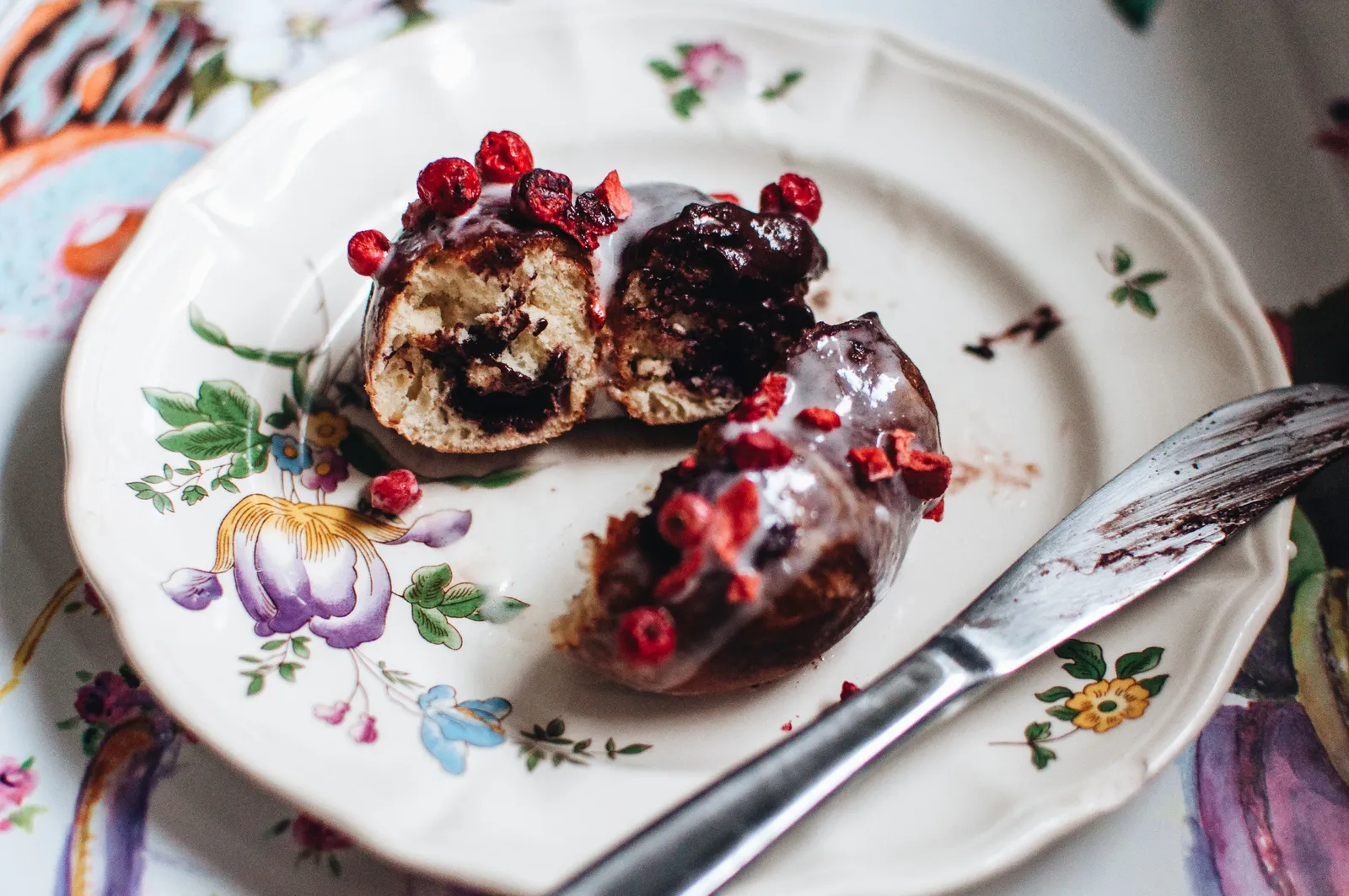 Ein angebissener Laugendonut mit dunkler Tahiniglasur und getrockneten roten Beeren auf einem weißen Porzellantellerchen mit Blumenmuster.