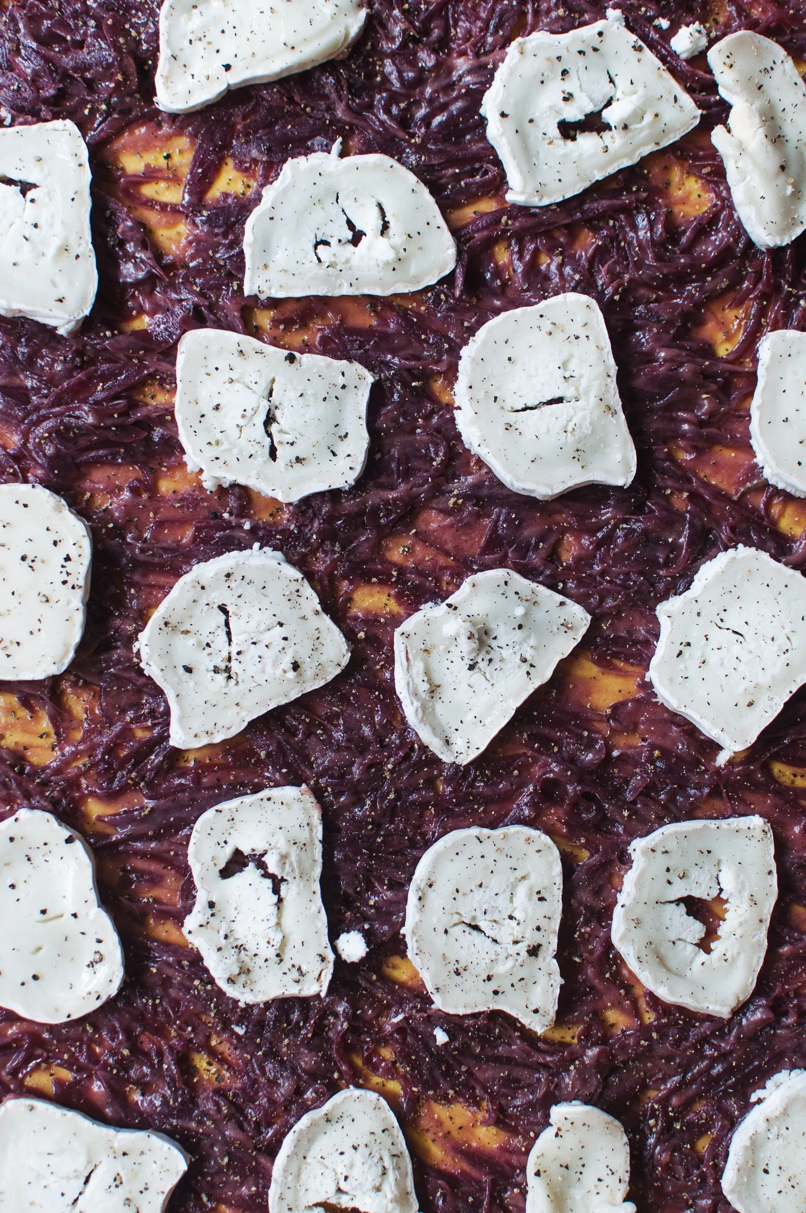 Ausgerollter Teig dicht belegt mit dunkelvioletten Rotweinzwiebeln, goldgelben Kürbistücken und unregelmäßigen Klecksen cremigem Ziegenkäse, mit schwarzem Pfeffer bestreut.