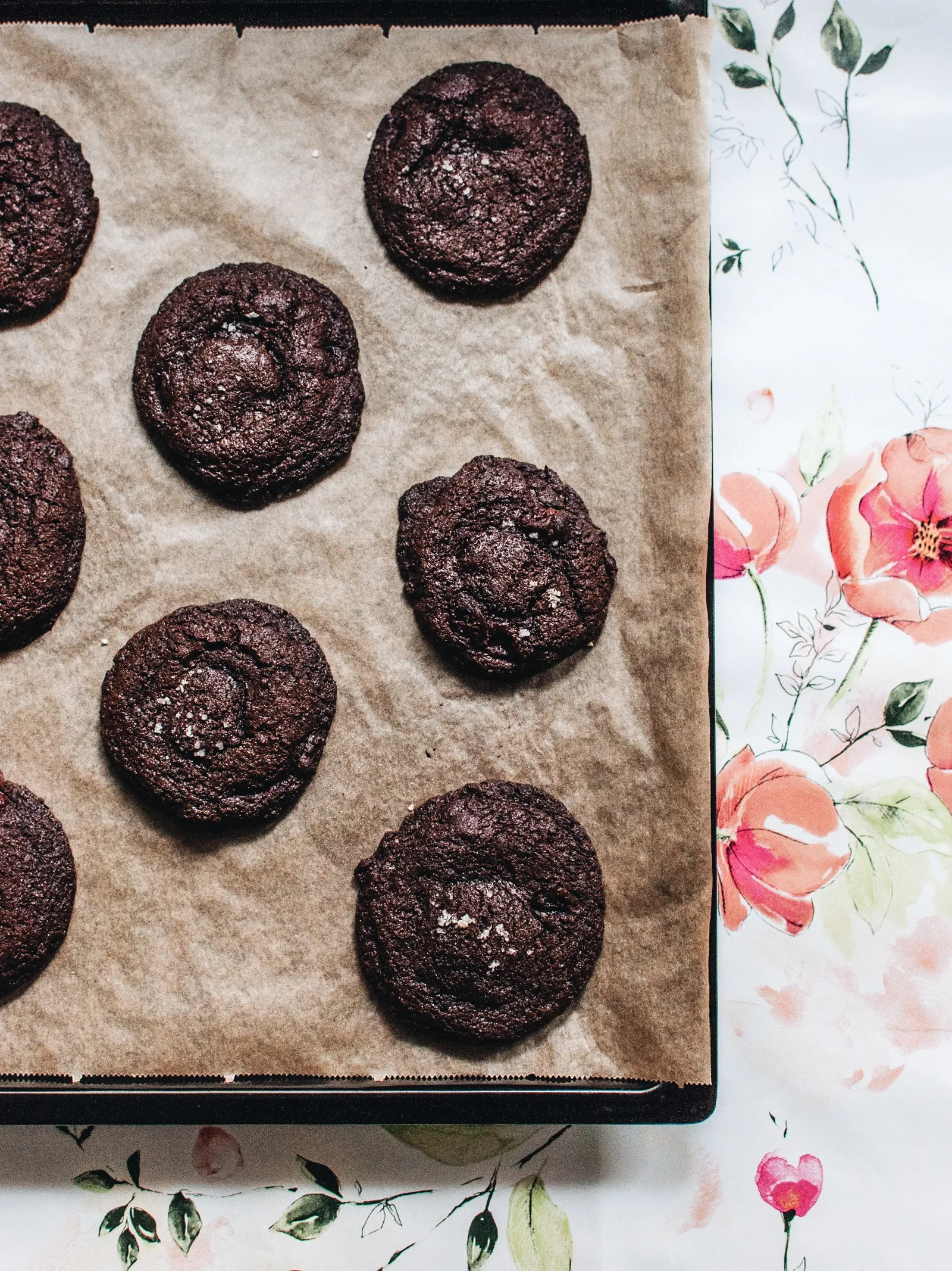 Dunkle Schokoladen-Cookies mit knuspriger Oberfläche und vereinzelten Salzflocken auf Backpapier, einer davon angebrochen.