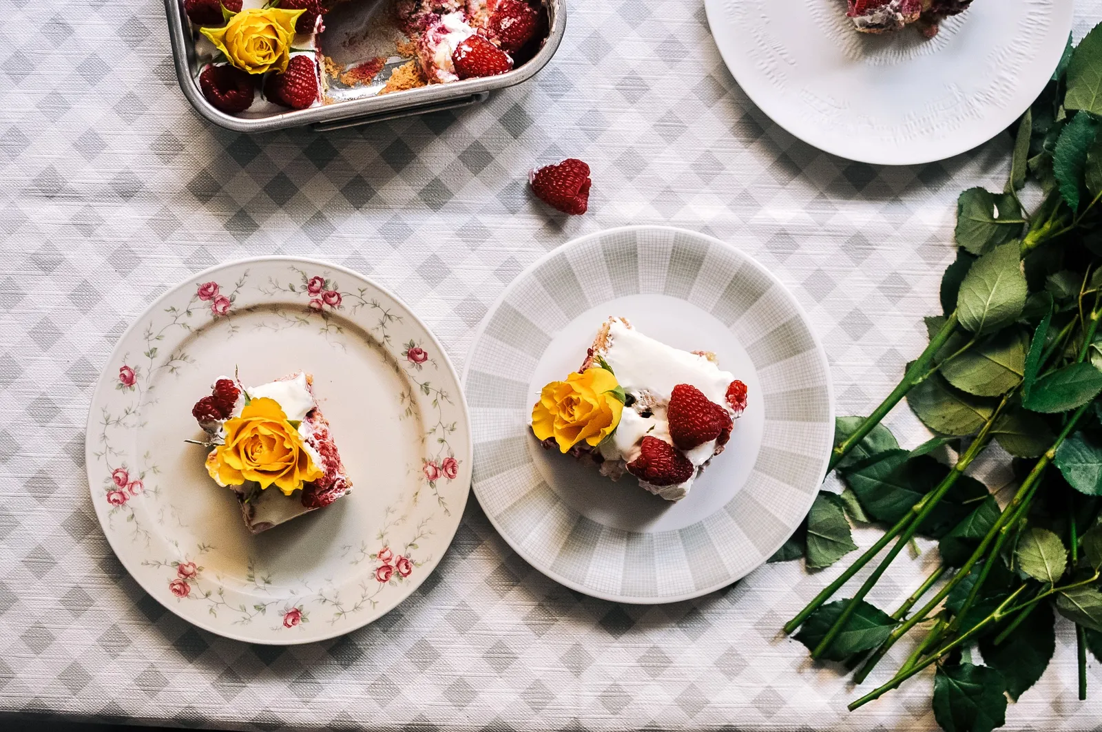 Ein Tres-Leches-Kuchen in einer rechteckigen Backform, dekoriert mit gelben Rosen, daneben Schälchen mit frischen Himbeeren und einer Kinderhand, die einen Rosenstiel hält – alles auf einem karierten Tischtuch.