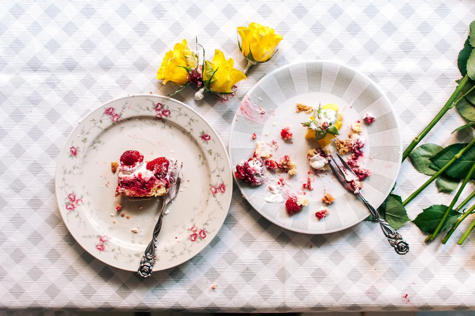 Eine Kuchengabel greift in einen Stück Tres-Leches-Cake mit Schlagsahne, Himbeeren und einer gelben Narzisse, daneben eine kleine Kinderhand – auf einem Karottiertischtuch mit zwei Tellern.